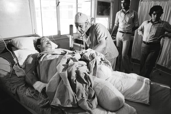 A black-and-white photograph of Bassam Shaka in a hospital bed being attended to by medical staff. His legs are amputated.