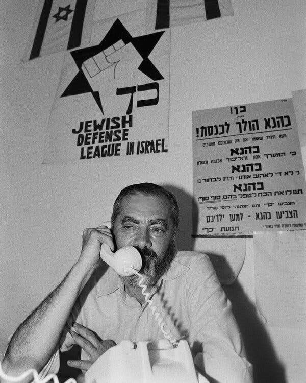 A black-and-white photograph of Meir Kahane on a phone with a “Jewish Defense League in Israel” sign behind him.
