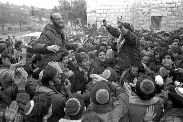 A black-and-white photograph of Rabbi Moshe Levinger being held aloft by a large group.